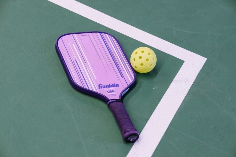 A purple pickleball paddle and yellow ball on a green court.